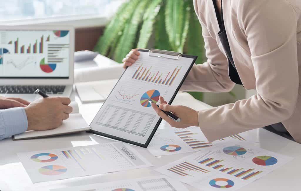 A business woman presenting a clipboard of graphs and pie chart to a business man behind a desk