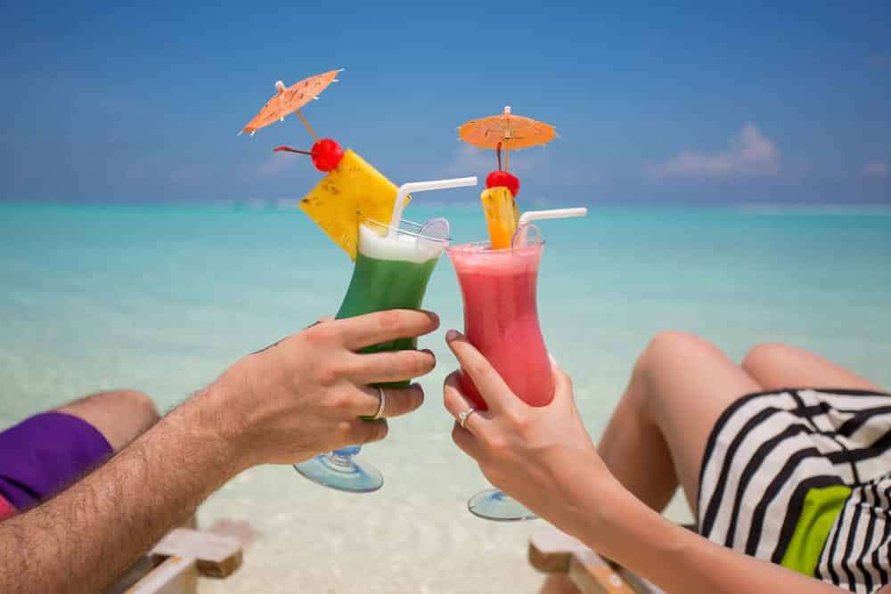 A man and woman clinking their umbrella drinks while laying down at a beach