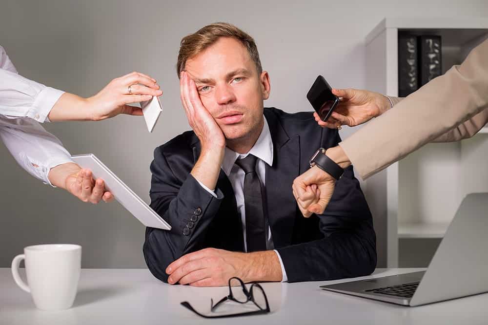 A stressed out looking business man at a desk with a laptop with four hands giving him phone and notes