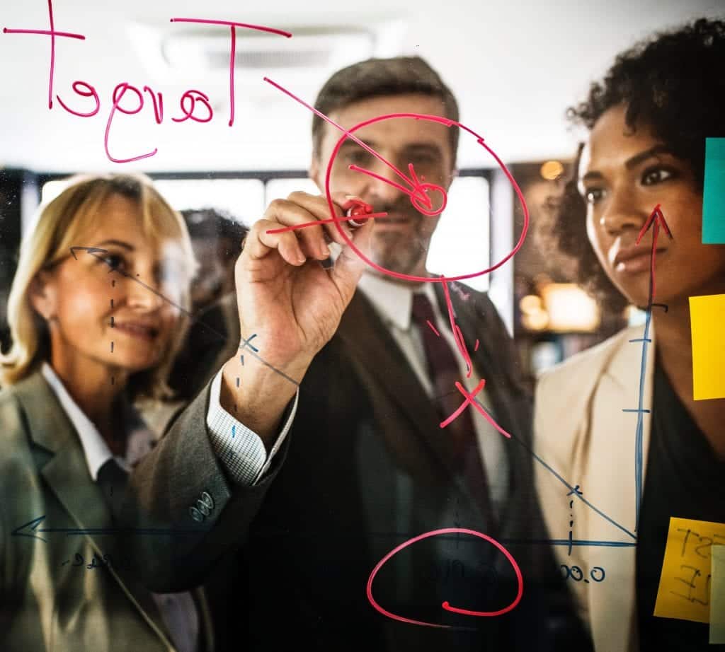 A man in a suit writing on a see through glass with dry erase marker while two women watch